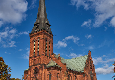 Eine Backsteinkirche mit hohem, spitzen Turm steht im Vordergrund, umgeben von herbstlichen Bäumen. Der Himmel ist blau mit einigen Wolken. Die Kirche hat große Fenster und einen Kupferdach. Der Eingang ist im gotischen Stil gestaltet.