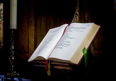 Ein aufgeschlagenes Buch, das auf einem Holzständer liegt. Die Seiten sind beschriftet, und es sind Lesezeichen in Rot und Grün sichtbar. Im Hintergrund steht eine Kerze.