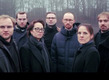 Eine Gruppe von sieben Menschen steht in einem bewölkten, nebligen Außenbereich. Sie tragen dunkle Kleidung und Blicken ernst in die Kamera. Der Hintergrund zeigt verschwommene Bäume, die eine ruhige, aber geheimnisvolle Atmosphäre schaffen.