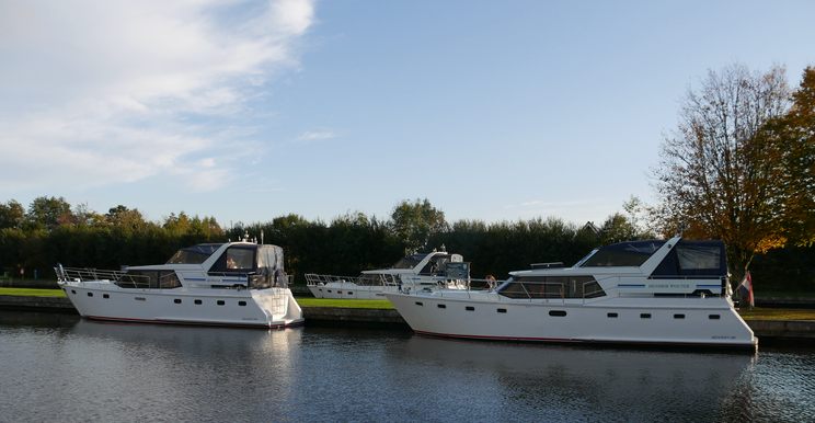 Giethoorn Hafen Drei weiße Boote liegenr in einem ruhigen Hafen. Im Hintergrund sind Bäume und ein klarer, blauer Himmel zu sehen. Die Szene vermittelt eine friedliche Atmosphäre.