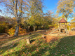 Ein sonniger Spielplatz im Herbst, umgeben von bunten laubtragenden Bäumen. Im Vordergrund steht ein Holzspielgerät mit Rutsche. Der Boden ist mit Herbstblättern bedeckt, und im Hintergrund ist eine kleine Holzhütte sichtbar.