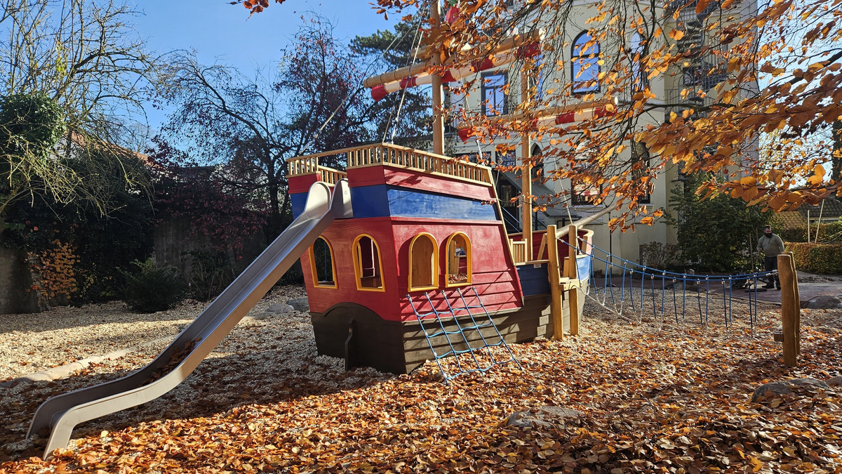 Eine bunte Spielplatzanlage in Form eines Piratenschiffs mit einer Rutsche und einer Kletterrampe. Der Bereich ist von herbstlichen Laub und Holzspänen umgeben. Im Hintergrund stehen Bäume und ein Gebäude.