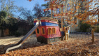 Eine bunte Spielplatzanlage in Form eines Piratenschiffs mit einer Rutsche und einer Kletterrampe. Der Bereich ist von herbstlichen Laub und Holzspänen umgeben. Im Hintergrund stehen Bäume und ein Gebäude.