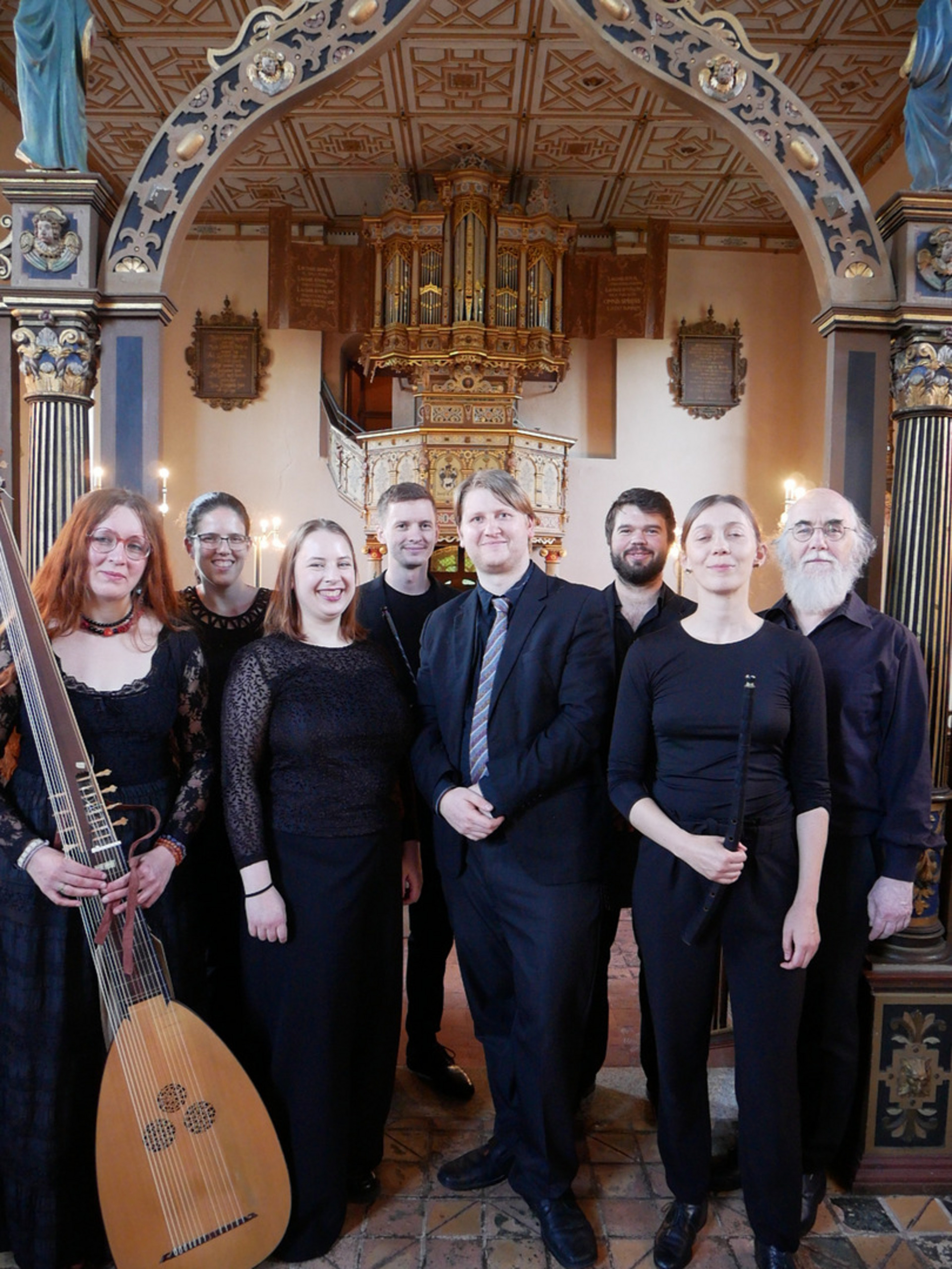 Eine Gruppe von acht Musikern steht in einer Kirche. Sie tragen formelle Kleidung und halten Instrumente in den Händen. Im Hintergrund ist eine ornate Orgel sichtbar. Die Atmosphäre ist festlich und musikalisch.