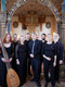Eine Gruppe von acht Musikern steht in einer Kirche. Sie tragen formelle Kleidung und halten Instrumente in den Händen. Im Hintergrund ist eine ornate Orgel sichtbar. Die Atmosphäre ist festlich und musikalisch.