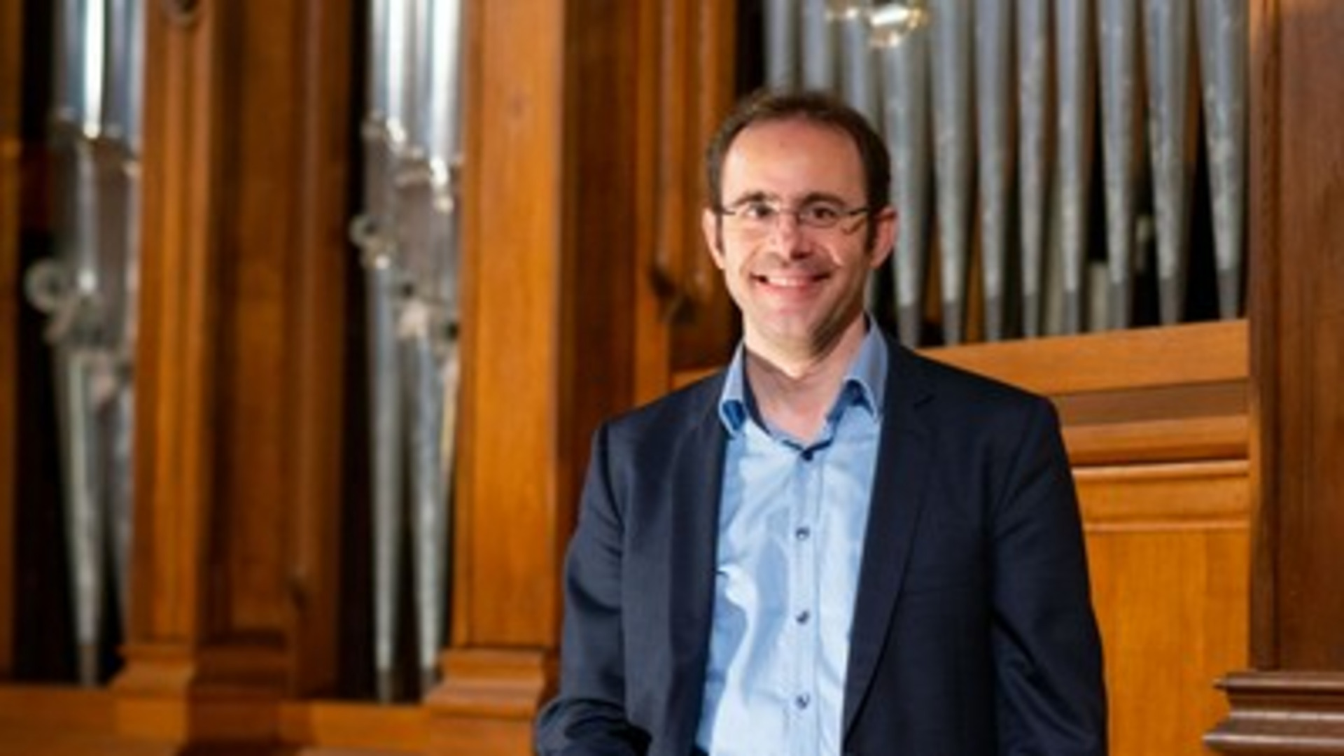 Ein lächelnder Mann steht vor einer Orgel aus Holz. Er trägt ein hellblaues Hemd und einen dunklen Anzug. Der Hintergrund zeigt die Pfeifen der Orgel, die in einer Kirche oder einem ähnlichen Raum platziert sind.