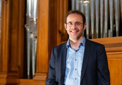 Ein lächelnder Mann steht vor einer Orgel aus Holz. Er trägt ein hellblaues Hemd und einen dunklen Anzug. Der Hintergrund zeigt die Pfeifen der Orgel, die in einer Kirche oder einem ähnlichen Raum platziert sind.