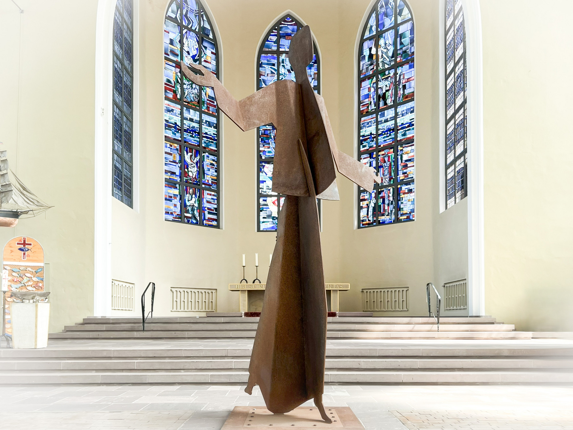 Eine große, stilisierte Metallstatue steht in einer Kirche. Die Figur hat ausgebreitete Arme und wirkt einladend. Bunte Glasfenster im Hintergrund zeigen religiöse Motive. Der Innenraum ist hell und schlicht gestaltet, mit einer Treppe und Kerzenhaltern im Mittelpunkt.