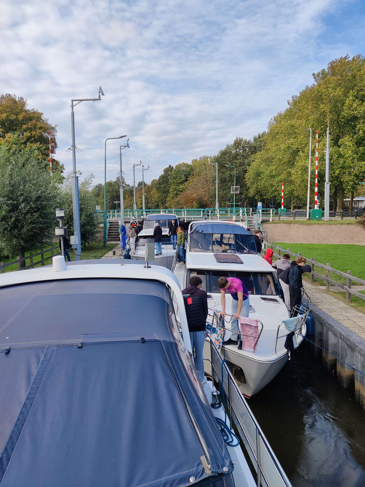 Marknessesluis Drei Boote liegen in einer engen Schleuse, während Menschen damit beschäftigt sind, sie zu Fendern und mit Leinen an Land festzuhalten. Einige Personen lächeln und unterhalten sich. Im Hintergrund sind Bäume und ein bewölkter Himmel zu sehen.