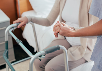 Eine Person sitzt auf der Bettkante und hält ihren Rollator fest. Eine weitere Person unterstützt sie dabei.