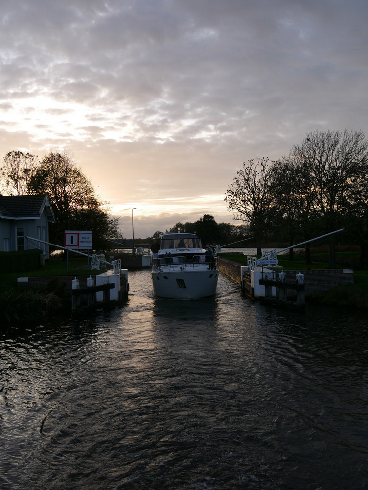 Kuinder von vorne Ein Boot fährt durch eine schmale Schleuse, links und rechts vom Boot sind die geöffneten Schleusentore. Das Boot ist von vorne zu sehen. Im Hintergrund sind Bäume und ein bewölkter Himmel sichtbar.