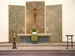 Der Altar in einer Kirche ist im Fokus. Über dem Altar hängt ein großes Kreuz mit einer Christusfigur. Auf dem Altar steht eine Blumenschale und eine Kerze. Im Hintergrund sind zwei große, graue Wandflächen zu sehen, die den Raum umrahmen. Rechts befindet sich ein Lesepult.