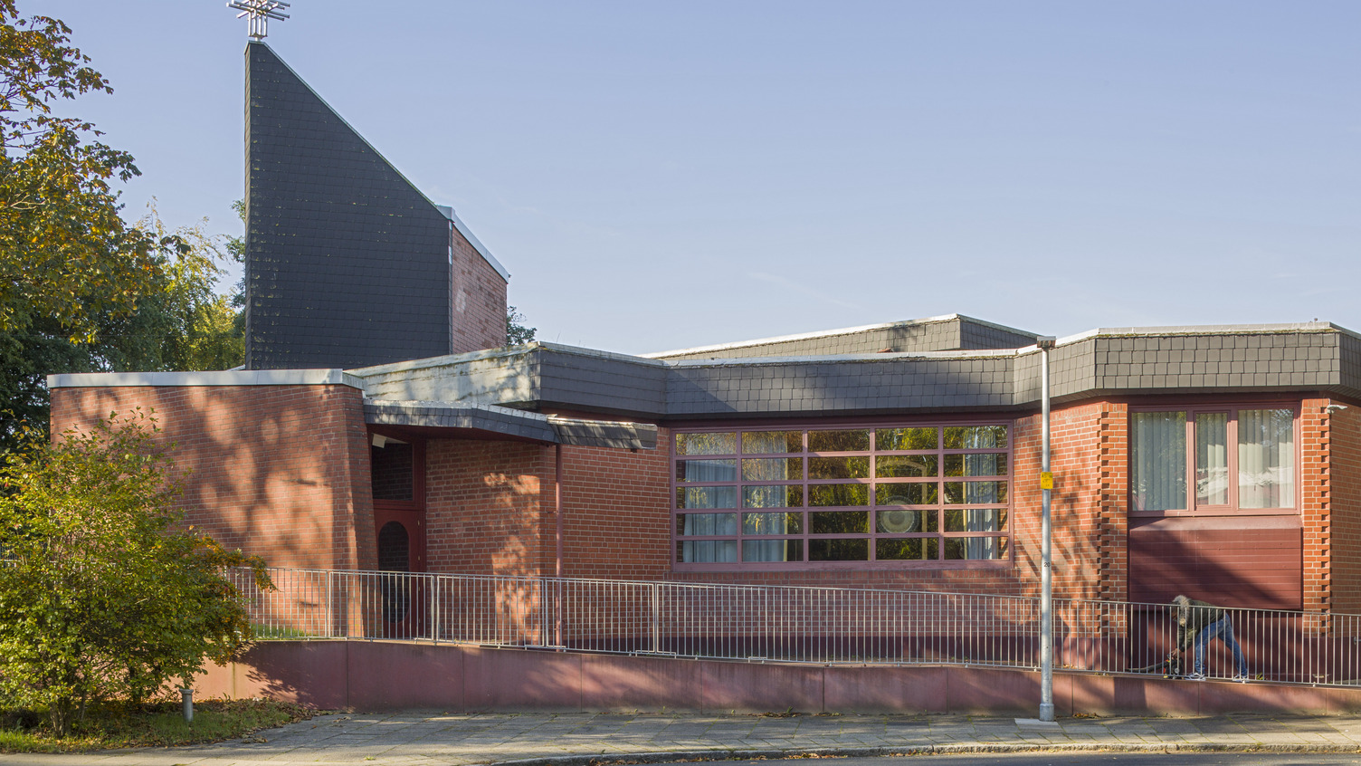 Ein modernes Gebäude mit roter Backsteinfassade und einem hohen, schrägen Dach. An der Spitze des Daches befindet sich ein metallisches Kreuz. Große Fenster lassen viel Tageslicht herein, und ein Weg führt zum Eingang. Bäume und ein gepflasterter Gehweg umgeben das Gebäude.