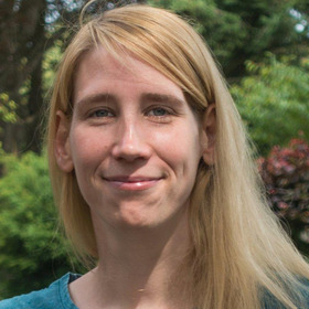 Eine lächelnde Frau mit langen, blonden Haaren und einem freundlichen Ausdruck steht im Freien. Der Hintergrund zeigt grüne Pflanzen und Sträucher. Die Person trägt ein einfaches, blaues Oberteil.