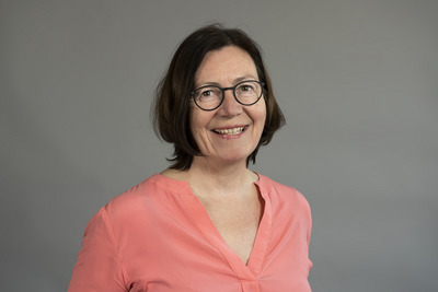 Porträt einer lächelnden Frau mit kurzen, braunen Haaren, die eine rosafarbene Bluse trägt. Sie trägt eine Brille und steht vor einem grauen Hintergrund.
