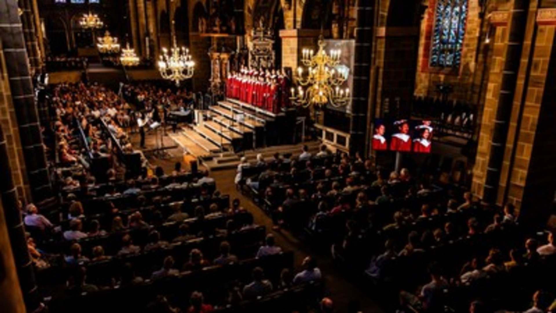 Ein großer Chor in rote Gewänder gekleidet singt auf einer erhöhten Bühne in einer Kirche. Vor dem Chor sitzt ein Publikum, das aufmerksam zuhört. Im Hintergrund sind beeindruckende Kirchenfenster und Kronleuchter zu sehen. Die Atmosphäre ist feierlich und musikalisch.