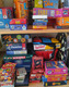 Das Bild zeigt ein Regal voller Spieleboxen. Darunter sind bekannte Spiele wie Uno, Skrabble, Tabu, und Twister. Die Boxen sind bunt und unterschiedlich gestapelt, was eine vielfältige Spielauswahl darstellt.