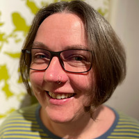 Porträt einer lächelnden Person mit kurzen, grauen Haaren und einer Brille. Sie trägt ein gestreiftes Oberteil in grünen und blauen Farben. Der Hintergrund zeigt ein lebhaftes, grünes Muster.