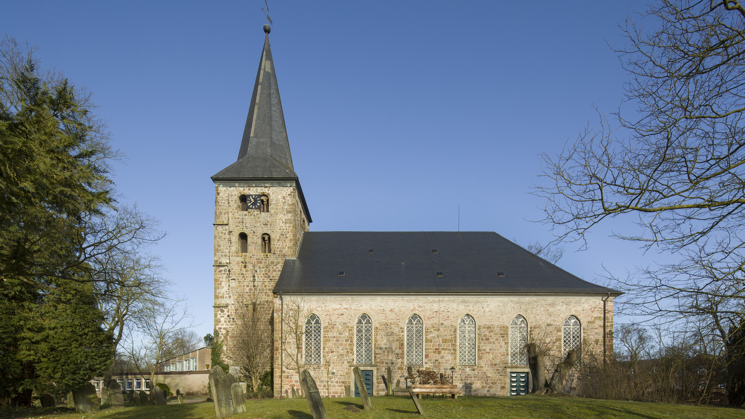 Die Bildbeschreibung zeigt eine Kirche mit einem hohen, spitzen Turm und charakteristischen Fenstern. Der Gebäudekörper ist aus helleren Ziegelsteinen, umgeben von einem ruhigen, grünen Rasen und einigen Grabsteinen im Vordergrund. Der Himmel ist klar und blau.