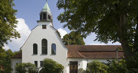 Eine weiße Kirche mit einem spitzen Dach und einem kleinen Glockenturm. Die Fassade hat große, oben abgerundete Fenster. Umgeben von Bäumen und Sträuchern, strahlt die Kirche eine ruhige Atmosphäre aus.