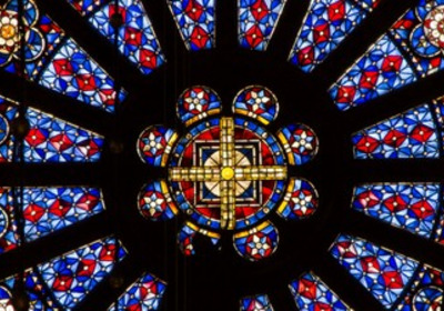 Das Bild zeigt ein farbenfrohes Kirchenfenster mit einem runden Motiv in der Mitte. Es ist mit roten und blauen Glasstücken sowie geometrischen Mustern verziert. Die zentrale Figur besteht aus goldenen und hellen Elementen, die die symmetrische Gestaltung betonen.