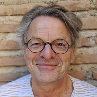 Ein älterer Mann mit grauem Haar und runden Brillenläsern lächelt freundlich vor einer steinernen Wand. Er trägt ein gestreiftes T-Shirt.