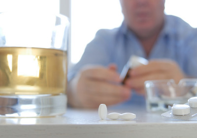Alkohol und Tabletten stehen auf einem Tisch, dahinter sitzt eine Person.