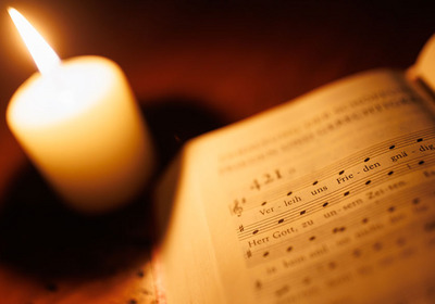 Das Foto zeigt ein aufgeschlagenes Gesangbuch und im Hintergrund eine leuchtende Kerze