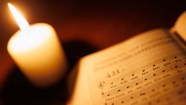 Das Foto zeigt ein aufgeschlagenes Gesangbuch und im Hintergrund eine leuchtende Kerze
