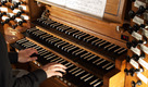 Ein Organist spielt auf einer Holzorgel. Die Hände berühren die schwarzen und weißen Tasten. Ein Notenblatt steht auf dem Spieltisch. Die Orgel hat verschiedene Register und Knöpfe zur Klangänderung.