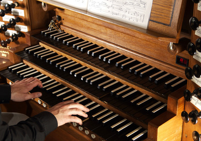 Ein Organist spielt auf einer Holzorgel. Die Hände berühren die schwarzen und weißen Tasten. Ein Notenblatt steht auf dem Spieltisch. Die Orgel hat verschiedene Register und Knöpfe zur Klangänderung.