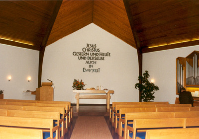 Der Innenraum einer Kirche mit Holzdecke und Reihen von Holzbänken. An der Wand steht in großen Buchstaben der Schriftzug: „Jesus Christus gestern und heute und derselbe auch in Ewigkeit“. Ein Tisch steht vorne mit religiösen Utensilien, und ein Organist sitzt an einer Orgel.