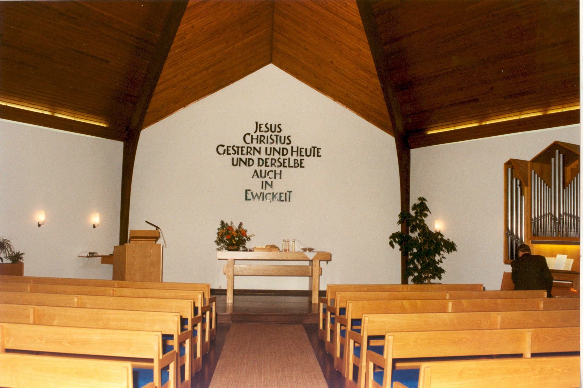 Der Innenraum einer Kirche mit Holzdecke und Reihen von Holzbänken. An der Wand steht in großen Buchstaben der Schriftzug: „Jesus Christus gestern und heute und derselbe auch in Ewigkeit“. Ein Tisch steht vorne mit religiösen Utensilien, und ein Organist sitzt an einer Orgel.