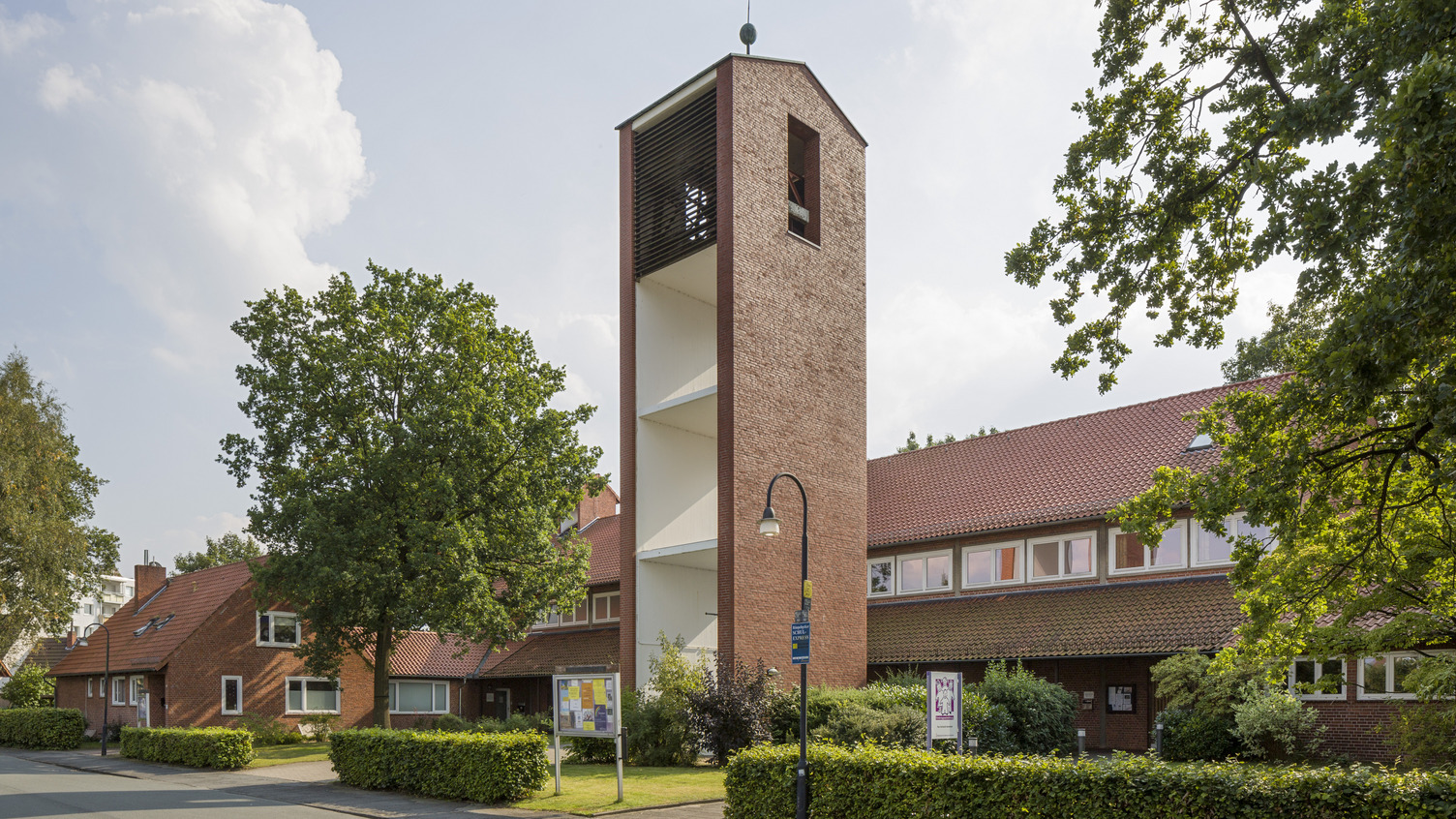 Das Bild zeigt ein rotes Ziegelgebäude mit einem hohen, schmalen Kirchturm, der ein Glockenspiel enthält. Vor dem Gebäude wachsen grüne Bäume und Sträucher. Die Umgebung ist gepflastert mit einer Straße und einem Gehweg. Der Himmel ist blau mit ein paar Wolken.