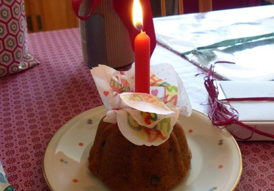 Ein kleiner, dekorativer Kuchen steht auf einem Teller, gekrönt von einer roten Kerze. Um die Kerze sind bunte Papierschnipsel, die Gratulationen aussprechen. Der Kuchen hat eine runde Form und wirkt festlich, symbolisch für eine Geburtstagsfeier oder einen besonderen Anlass.