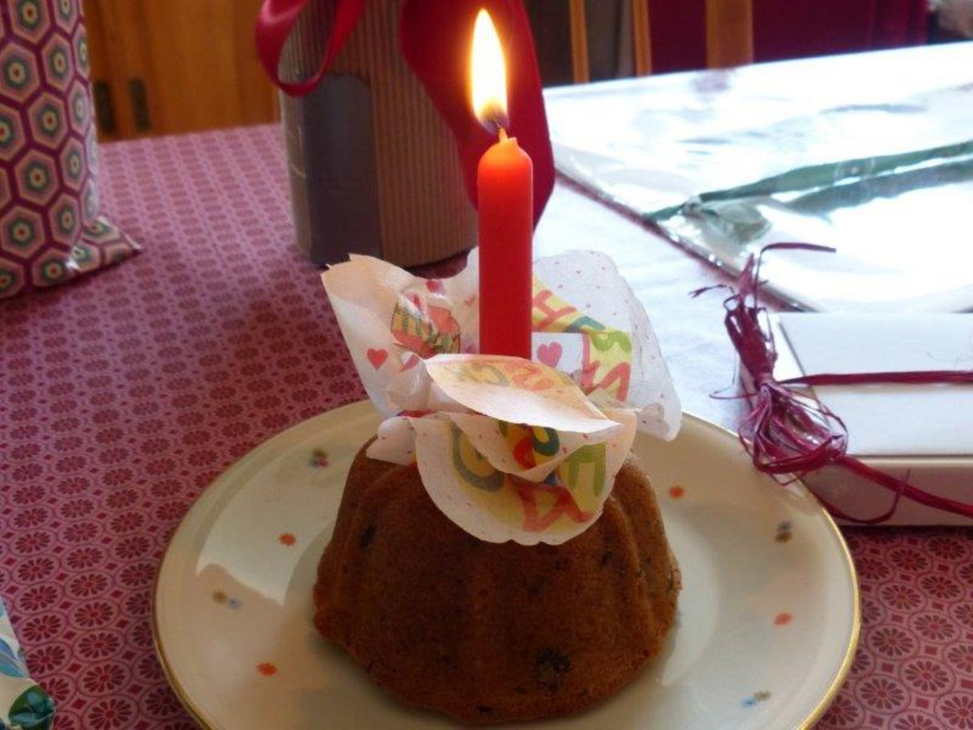 Ein kleiner, dekorativer Kuchen steht auf einem Teller, gekrönt von einer roten Kerze. Um die Kerze sind bunte Papierschnipsel, die Gratulationen aussprechen. Der Kuchen hat eine runde Form und wirkt festlich, symbolisch für eine Geburtstagsfeier oder einen besonderen Anlass.