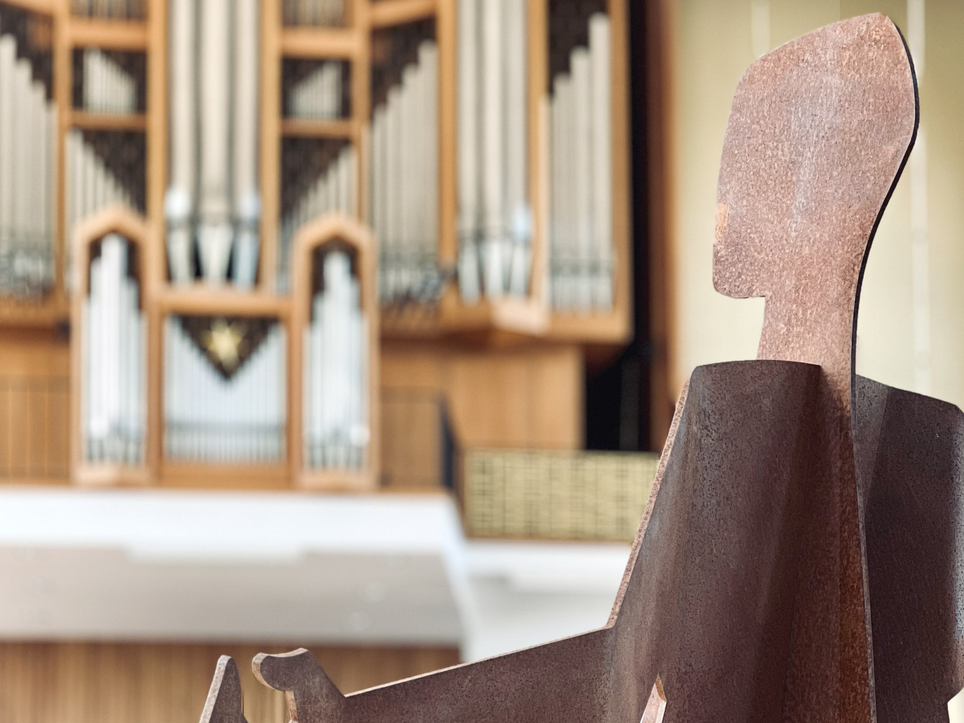 Die Abbildung zeigt eine stilisierte Skulptur einer sitzenden Figur im Vordergrund, vor einer großen Orgel im Hintergrund. Die Statue ist aus einem roströtlichen Material gefertigt und hebt sich von der hellen Umgebung ab.