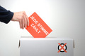 Die Illustration zeigt eine Hand, die einen Stimmzttel in eine Wahlurne wirft