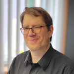 Ein Mann mit Brille trägt ein schwarzes Hemd und steht lächelnd in einem hellen Raum mit Fensterjalousien. Der Hintergrund zeigt die Konturen von Möbeln und Pflanzen, die für eine Büroumgebung typisch sind.