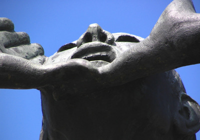 Eine Skulptur, die ein Gesicht von unten darstellt. Der Ausdruck wirkt entschlossen und kraftvoll, mit angespannten Lippen und erhobenem Kinn. Die Hände halten das Gesicht sanft, was eine Verbindung oder Unterstützung symbolisieren könnte. Der Hintergrund ist klar und blau.