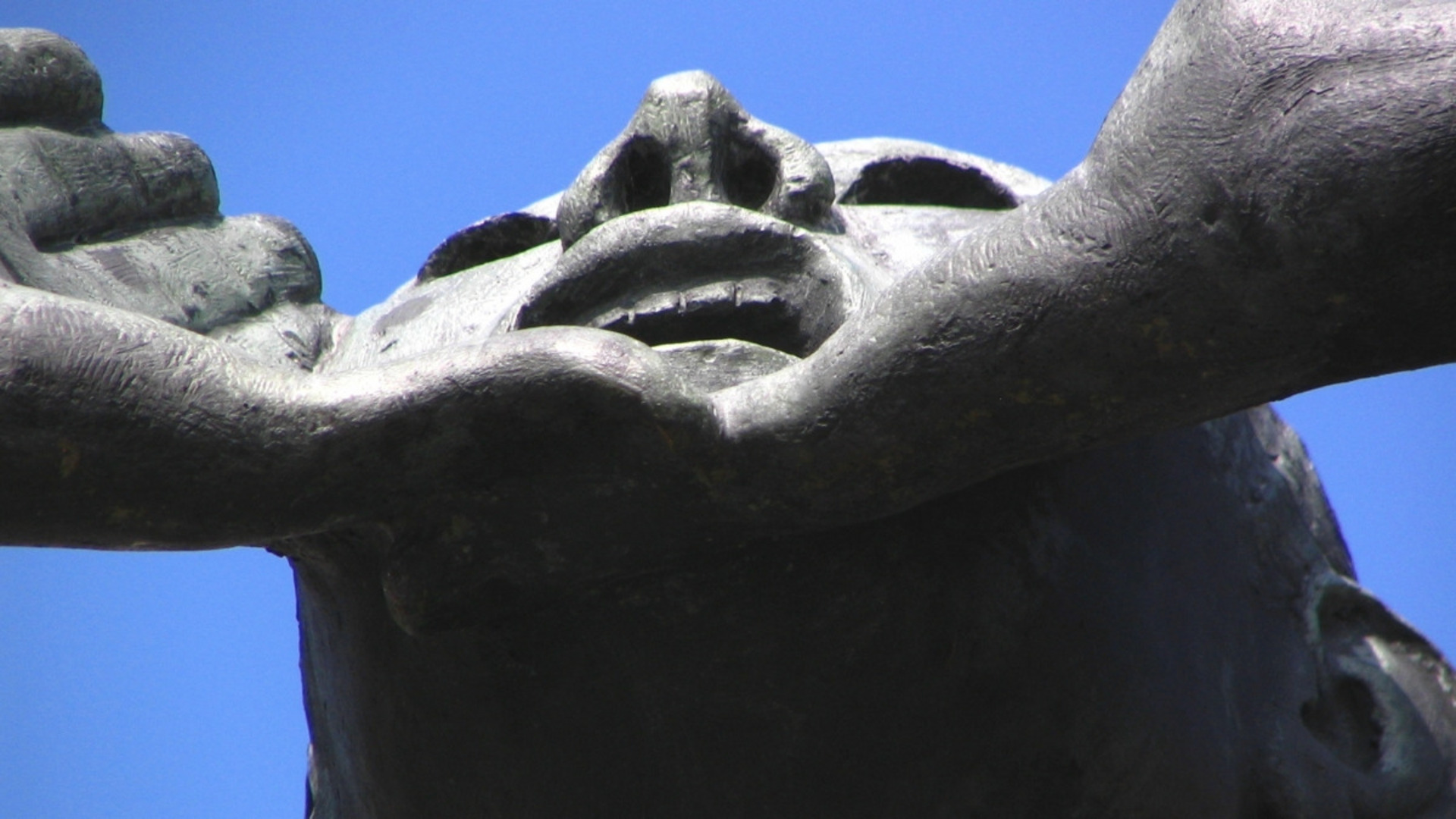 Eine Skulptur, die ein Gesicht von unten darstellt. Der Ausdruck wirkt entschlossen und kraftvoll, mit angespannten Lippen und erhobenem Kinn. Die Hände halten das Gesicht sanft, was eine Verbindung oder Unterstützung symbolisieren könnte. Der Hintergrund ist klar und blau.
