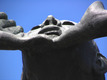Eine Skulptur, die ein Gesicht von unten darstellt. Der Ausdruck wirkt entschlossen und kraftvoll, mit angespannten Lippen und erhobenem Kinn. Die Hände halten das Gesicht sanft, was eine Verbindung oder Unterstützung symbolisieren könnte. Der Hintergrund ist klar und blau.