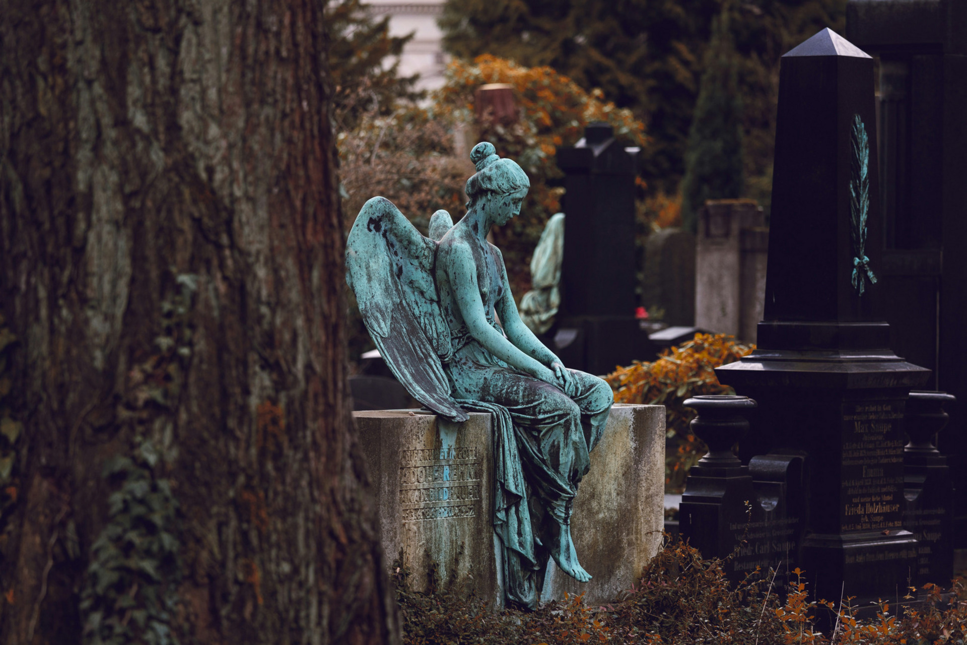 Eine patinierte Engelstatue sitzt melancholisch auf einem Grabstein, umgeben von weiteren Grabmalen und herbstlichen Pflanzen. Die Statue hat große Flügel und trägt ein schlichtes Gewand, während ihre Haltung nachdenklich wirkt.