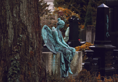 Eine patinierte Engelstatue sitzt melancholisch auf einem Grabstein, umgeben von weiteren Grabmalen und herbstlichen Pflanzen. Die Statue hat große Flügel und trägt ein schlichtes Gewand, während ihre Haltung nachdenklich wirkt.
