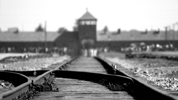 Einfahrt des Konzentrationslager Auschwitz-Birkenau; schwarz-weiß-Aufnahme; im Vordergrund Schienen, im Hintergrund unscharf das markante Tor mit dem Wachturm.