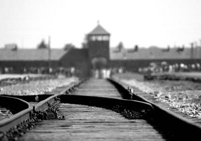 Einfahrt des Konzentrationslager Auschwitz-Birkenau; schwarz-weiß-Aufnahme; im Vordergrund Schienen, im Hintergrund unscharf das markante Tor mit dem Wachturm.
