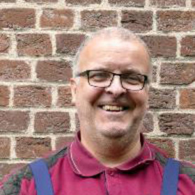 Ein lächelnder Mann mit Brille trägt ein rotes Poloshirt und hat einen freundlichen Ausdruck. Er steht vor einer Backsteinmauer, die als Hintergrund dient.