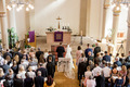 Zu sehen ist ein voller Kirchsaal, festlich eingerichtet für eine Hochzeit. Die anwesenden Gäste und das Brautpaar haben sich erhoben, um gemeinsam zu beten/ singen.