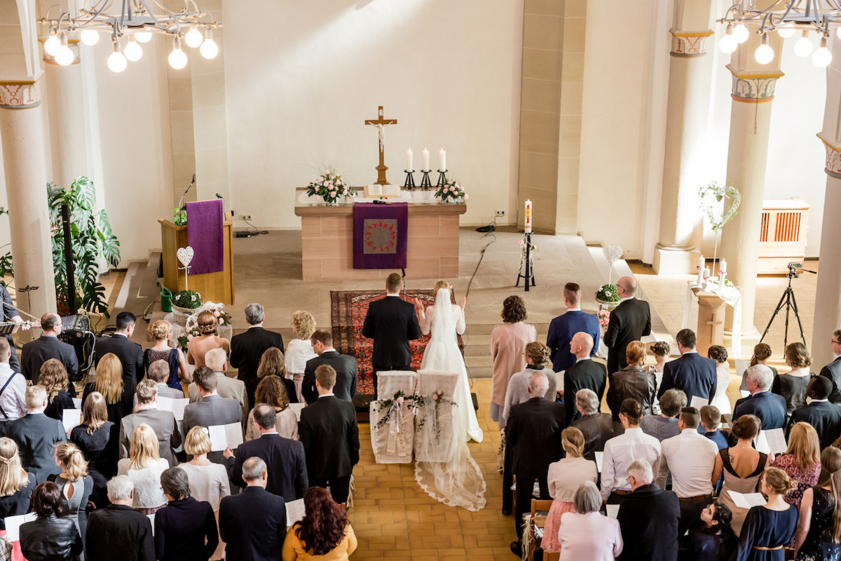 Hochzeitsgebet Zu sehen ist ein voller Kirchsaal, festlich eingerichtet für eine Hochzeit. Die anwesenden Gäste und das Brautpaar haben sich erhoben, um gemeinsam zu beten/ singen.
