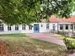 Ein helles Gebäude mit weißen Wänden und blauen Türen, umgeben von grünem Gras und Laubbäumen. Der Eingangsbereich ist gepflastert, und mehrere Fenster sind sichtbar. Im Hintergrund sieht man einen modernen Anbau. Das Gesamtbild vermittelt eine einladende Atmosphäre.